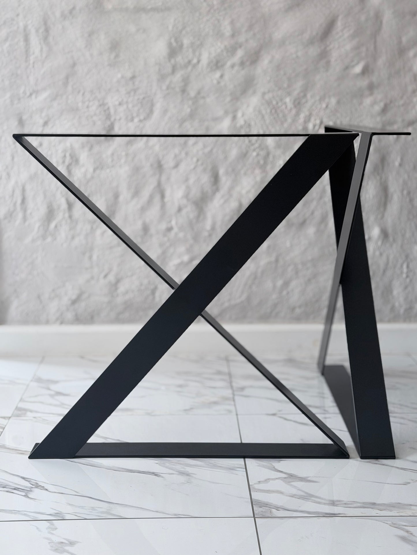 A modern black metal table base with a geometric X-shaped triangular support structure and a clear glass tabletop, set against a textured white wall and light marble floor.