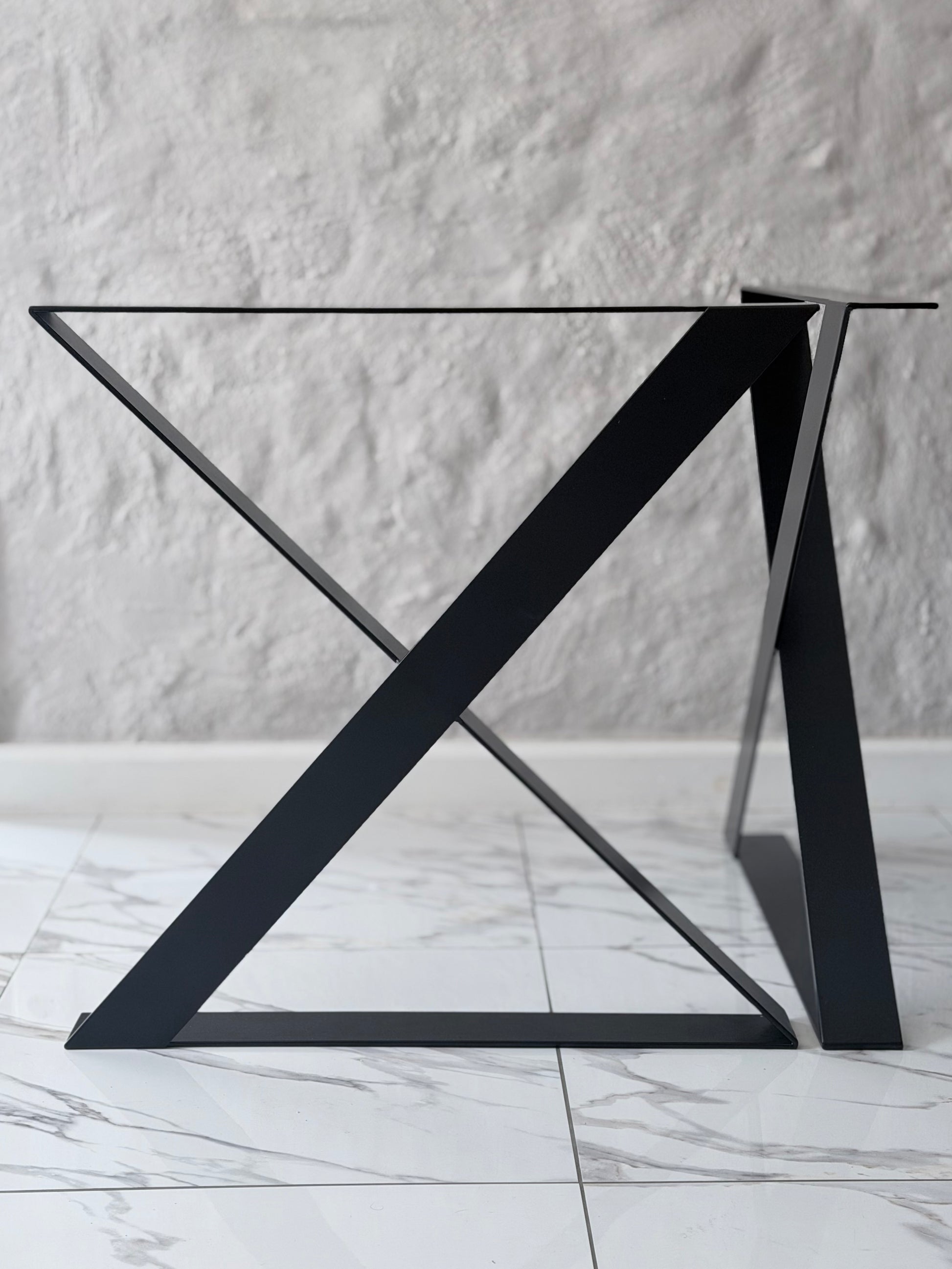 A modern black metal table base with a geometric X-shaped triangular support structure and a clear glass tabletop, set against a textured white wall and light marble floor.