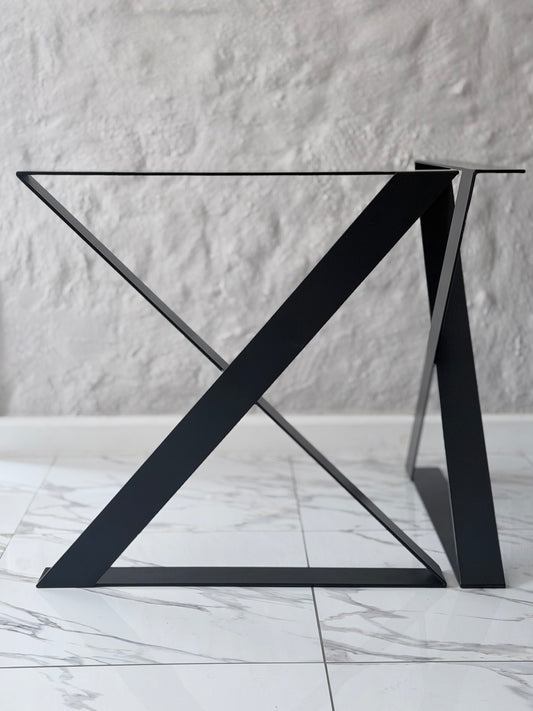 A modern black metal table base with a geometric X-shaped triangular support structure and a clear glass tabletop, set against a textured white wall and light marble floor.